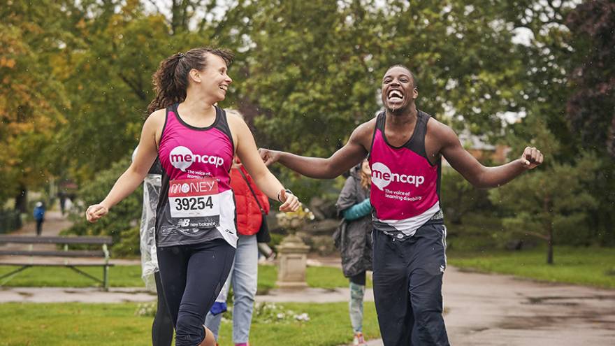 Man and woman wearing Mencap running vests, running together in park.