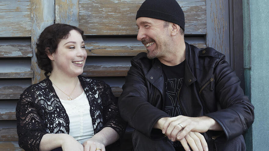 Man, The Edge, sat on step next to woman, Ciara. They are looking at each other and smiling.