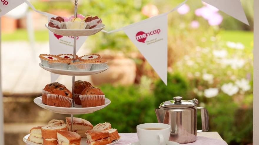 Photograph of the top of a table in a garden. The table has a table cloth with a cake stand full of scones and pastries, with a tea cup and saucer next to it. Mencap branded bunting hangs above the table.