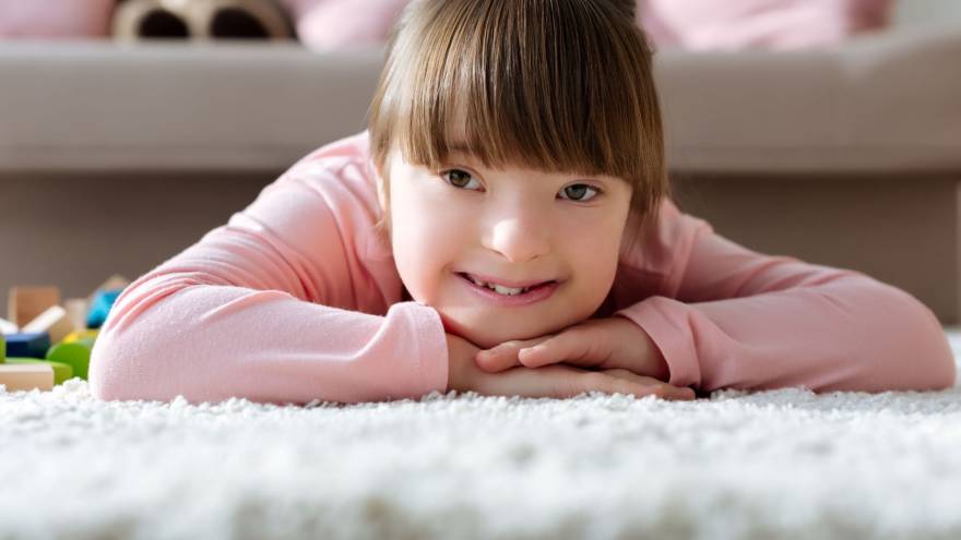 Child with downs syndrome lying on a rug facing the camera with head on folded arms and smiling