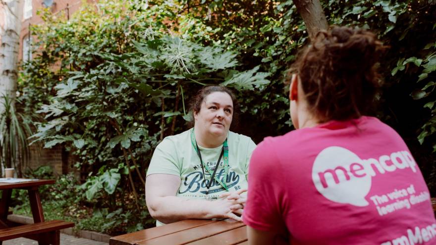 A woman talking to a Mencap support worker sitting at a picnic bench by a hedge