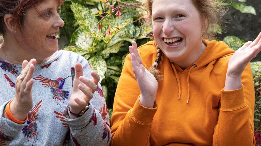 A girl with learning disabilities sitting outside next to a woman. They are both smiling and clapping their hands.