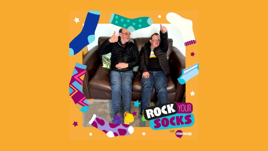 Two men, one with Down's Syndrome sitting on a brown leather sofa smiling with their thumbs in the air