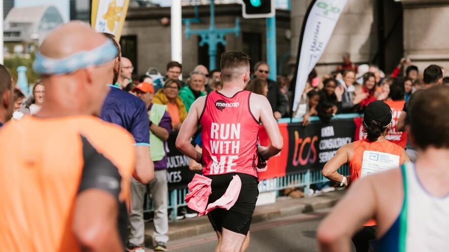The back of a mail marathon runner wearing a Mencap running vest on a street running a marathon course with other runners.