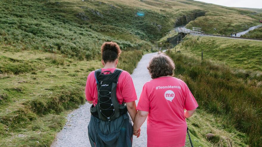 Two women walking away from the camera and up a path surrounded by moorland. Both are wearing pink Mencap Tshirts and one has a ruck sack.