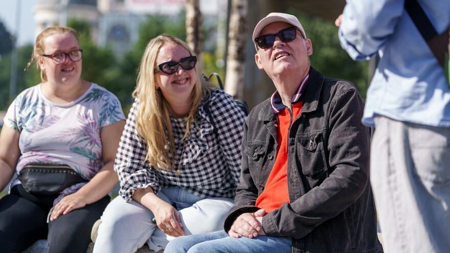 Three people sitting on a wall looking up to a man standing in front of them talking