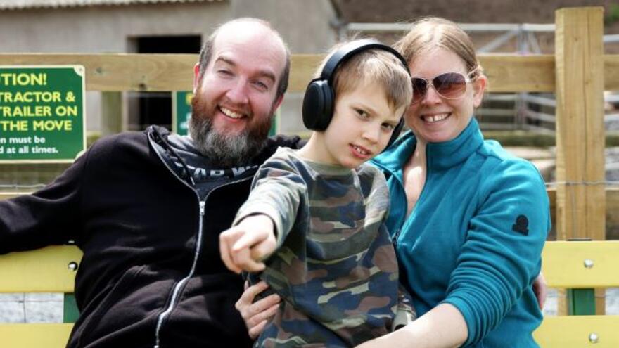 A mother and father either side of a boy with ear phones who is pointing at the camera. Behind them is a fence in a children's zoo