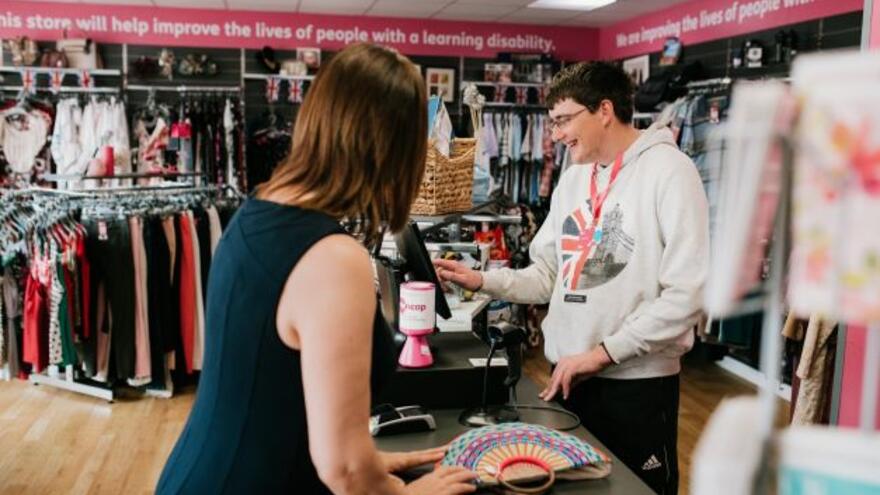 A man serving a female customer whilst using a till in a Mencap shop 