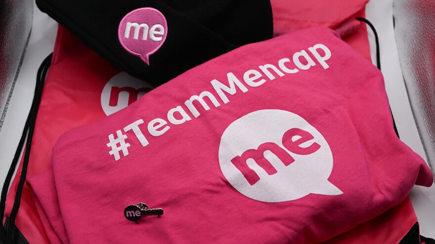 A photograph of a Mencap pin badge on top of a Mencap T-shirt next to a Mencap beanie hat
