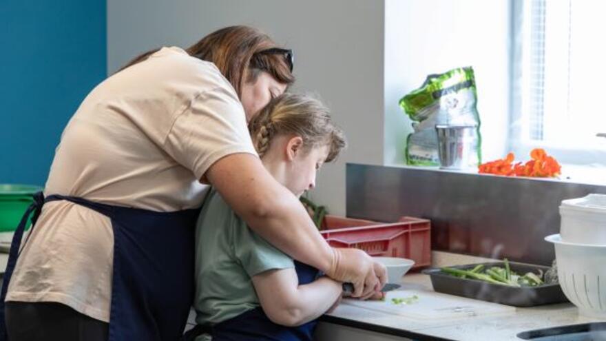 A woman helps another woman to chop vegetables in a kitchen