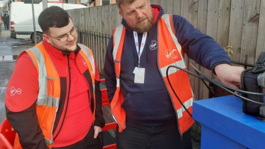 A man is teaching an intern how to use some equipment. Both are wearing high visibility vests