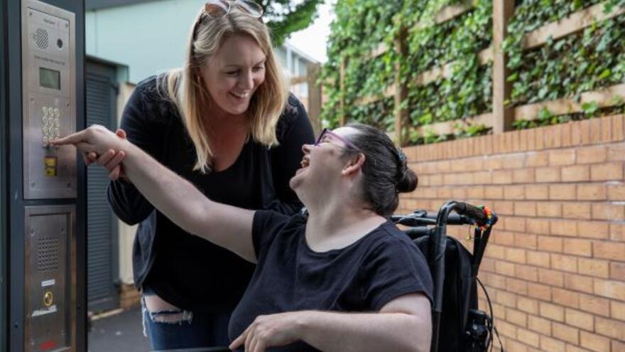 A woman in a wheelchair is putting some money into a car park pay machine and her supporter is smiling at her