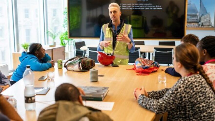 A man in a high visibility vest is talking to pupils in a classroom in front of a blackboard