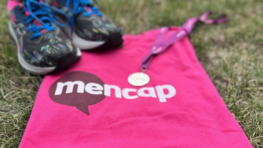 A pair of running shoes is on the grass alongside a Mencap medal and a Mencap T-shirt