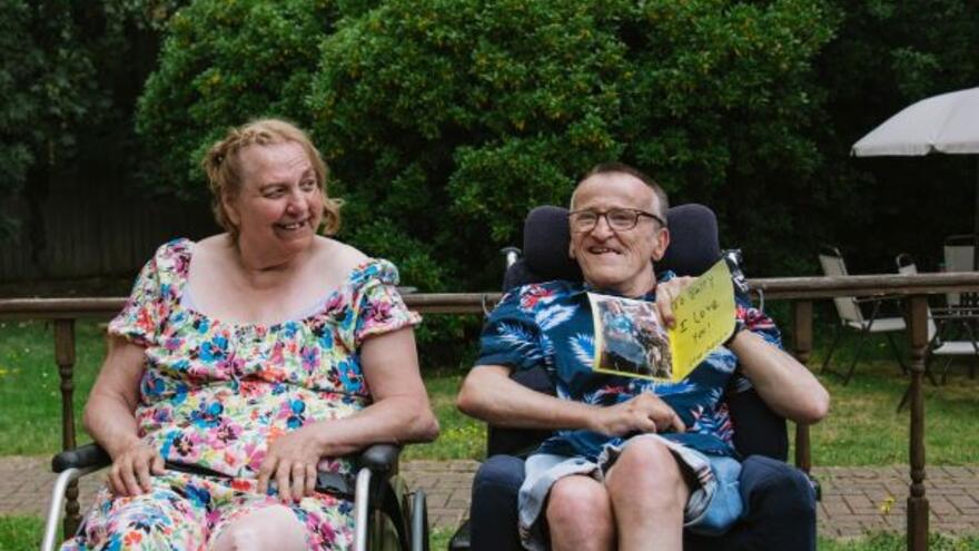 Betty and Barry are in a garden sitting in their wheelchairs next to each other. Barry is holding up a card which says To Barry I love you love Betty