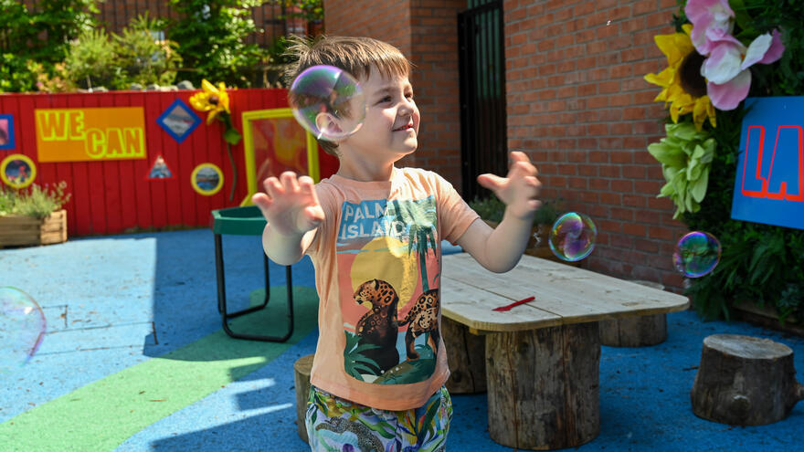 A young boy is waving his arms at some bubbles which are blowing in the wind in a playground
