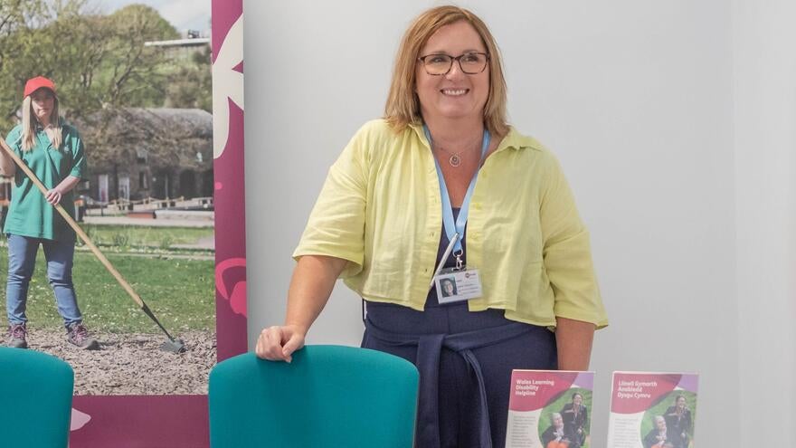 A woman with an ID badge around her neck, standing behind a desk with Mencap leaflets