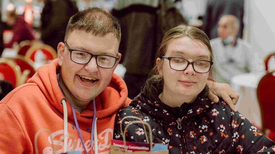 A young man and woman are sat at a table in a community centre smiling at the camera