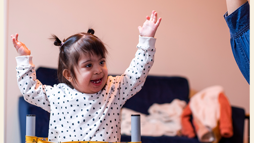 A young child in a harness holds her hands up and smiles