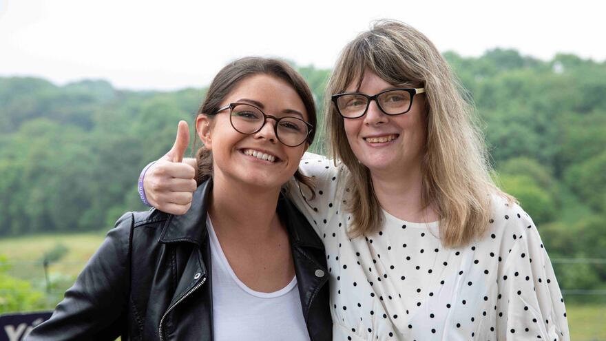 Two women are standing against a fence in the countryside. One has her arm around the other and her thumb up