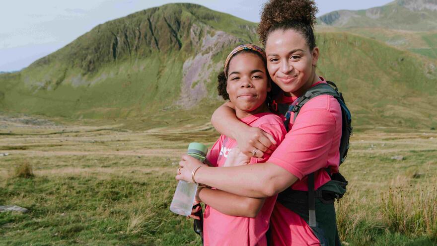 Two young women in Mencap T-shirts hugging at the foot of a mountain