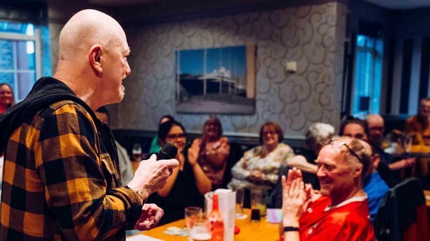 A man with a microphone is standing in front of a room full of people who are sitting at tables clapping