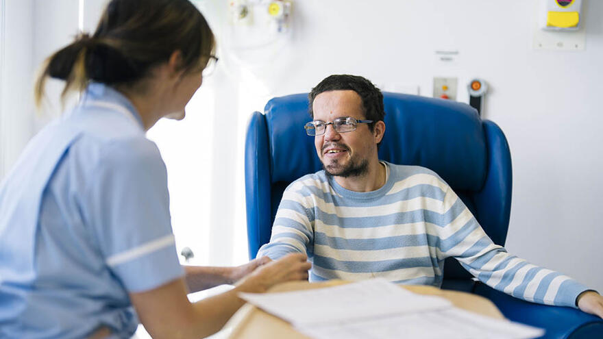 A man in a striped jumper, sits in a chair and smiles as a female nurse checks his pulse.