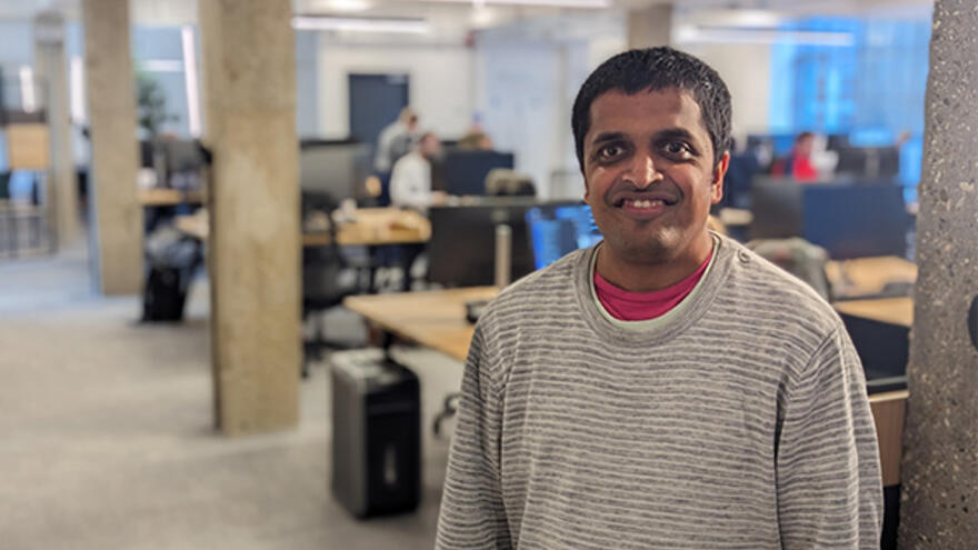 Vijay standing in office with people working on computers at desks behind him