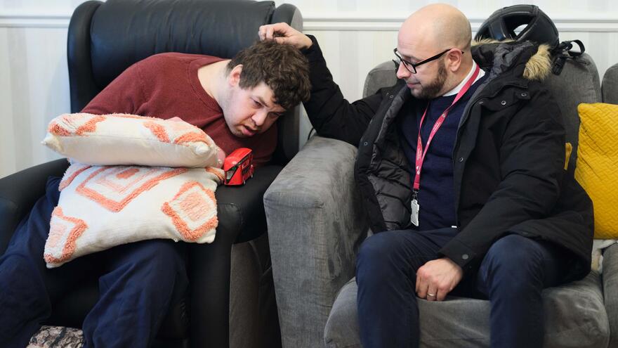 A support worker is sat on a comfy chair next to a young man who has a learning disability. The support worker is carefully stroking his head.