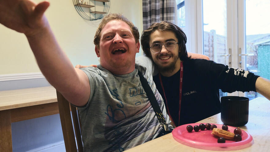 A support worker is sitting next to someone he cares for at a table smiling.