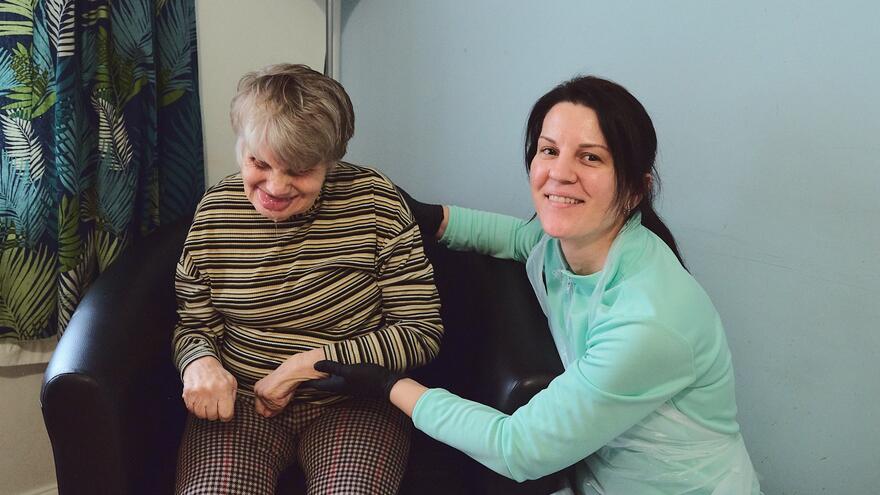 A support worker is crouched down next to the lady she cares for who is sitting on a chair