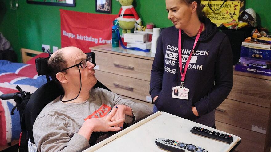A support worker talks to a man who is sitting at a desk in his bedroom