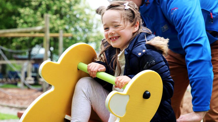 A man pushes his daughter on a rocking horse in a playground