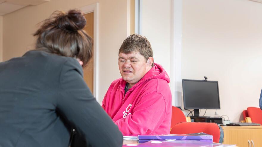Two people at a desk in an office. One person is writing things down and the other is talking to them from behind the desk.