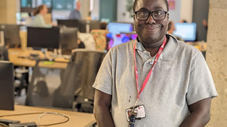 Richard in a grey polo shirt and glasses, standing in an office with people working on computers on desks behind him.