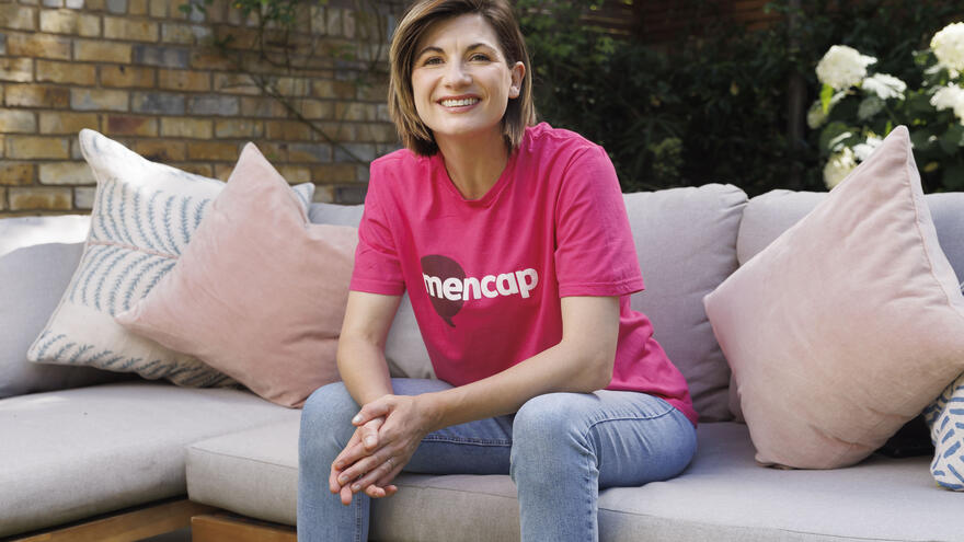 Actress Jodie Whittaker sitting on a sofa wearing a Mencap t-shirt smiling at the camera