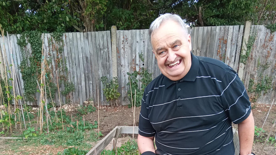 A man with grey hair and a dark shirt standing on an allotment with a wooden fence behind him