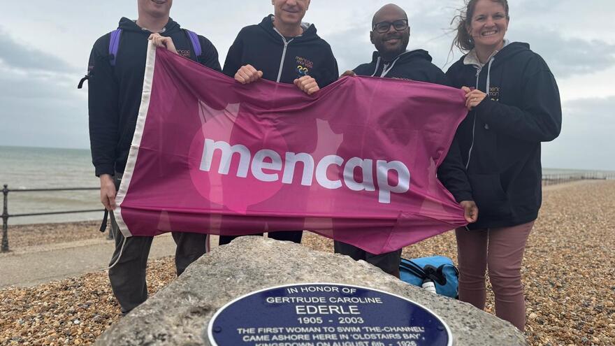 4 adults lined up at the shore holding a banner that says mencap