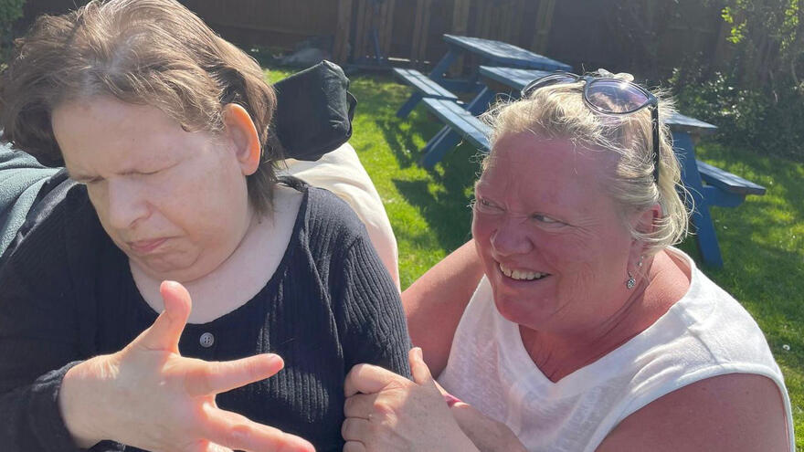 Two women next to each other in a garden. Caroline on the left has a black top and is sitting in a wheelchair. Her sister, Julie, on the right, wears white and is looking at Caroline and smiling.