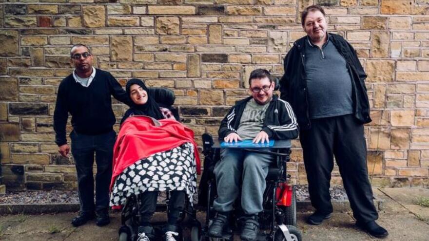 A group of four people with learning disabilities in a line, in front of a wall, facing the camera