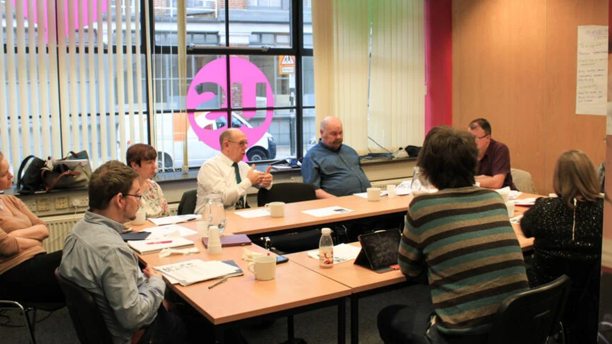A group of people sat around a U shaped meeting table talking