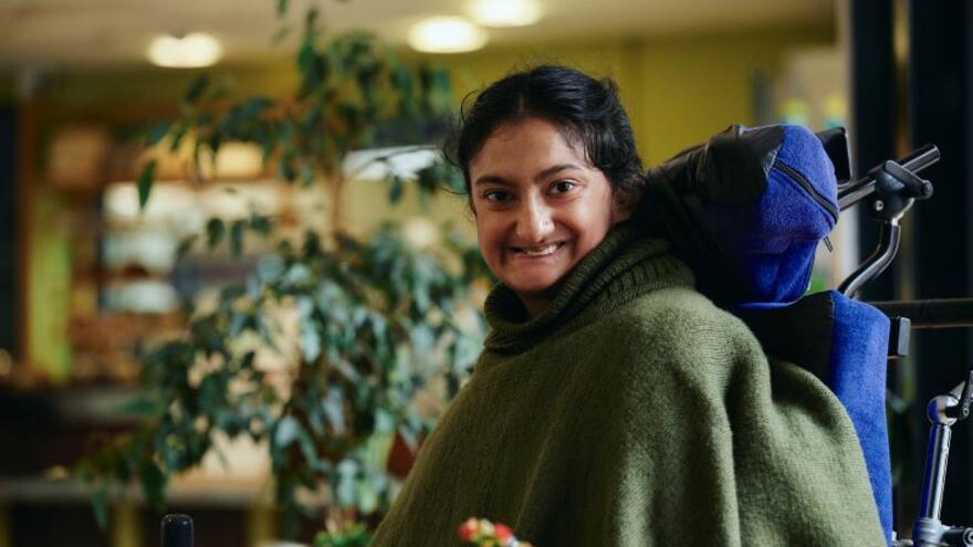 Rahima in a cafe sitting at a table in her wheelchair smiling at the camera