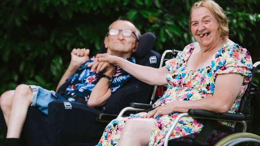 A man and a woman both in wheelchairs are holding hands.