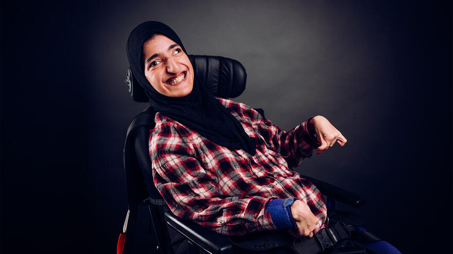A woman with a red checked short and a headscarf smiling at the camera. She is sitting in a wheelchait in front of a dark nackground