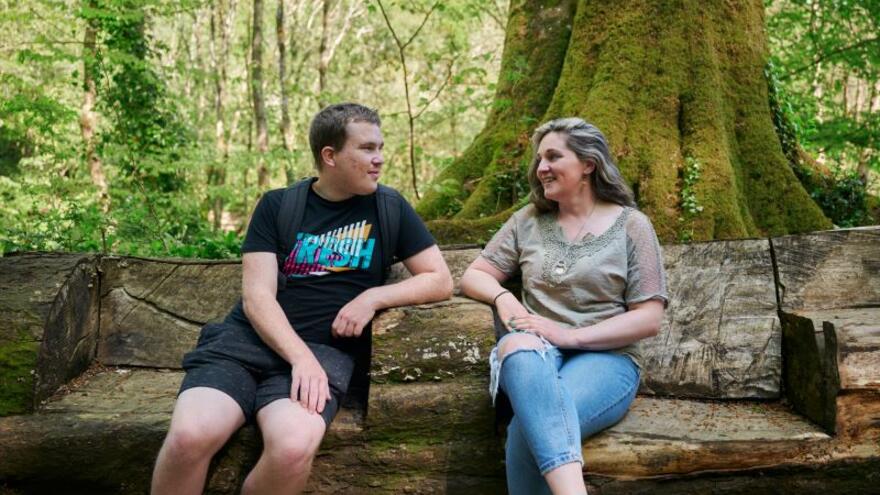 A man and a woman sitting on a bench made from a fallen tree, talking in the woods