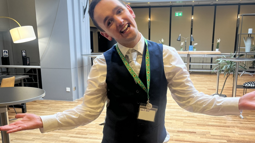 Supported internship learner, Xavier in a smart uniform standing in a hotel lobby. His hands are outstretched and he is smiling.