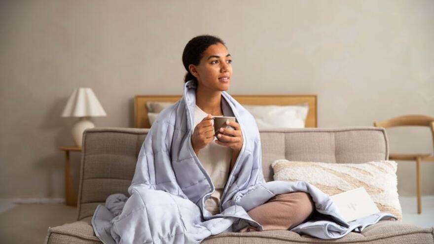 A young woman sitting on bed with a hot drink and a blanket around her