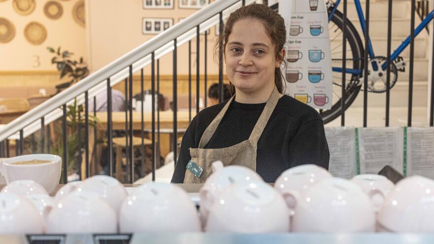 Supported internship learner, Maes stands behind a coffee machine at Fair Shot Café. There are lots of coffee cups stacked in front of her.