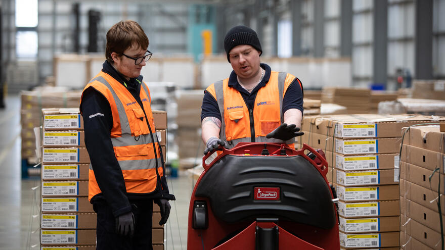 Two men in a warehouse wearing high-vis jackets. One of the men is showing the other how to use a piece of machinery.
