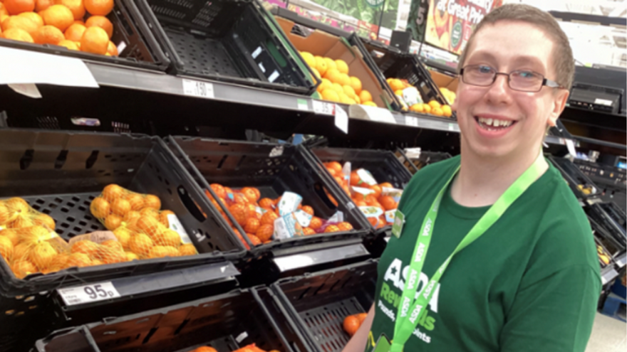 A supported internship learner in Asda uniform, smiling next to some of the produce.